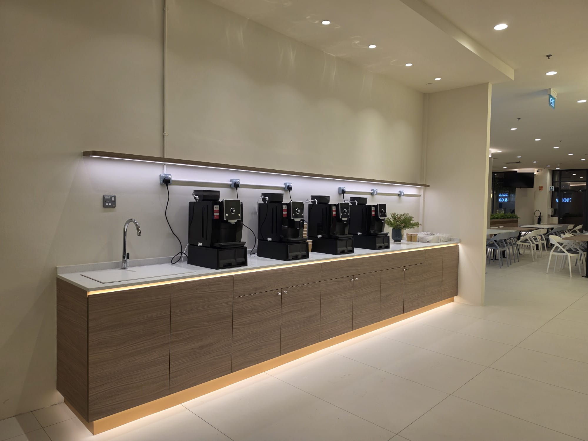 A pantry with timber cabinetry, a white countertop, integrated lighting and a row of coffee machines.